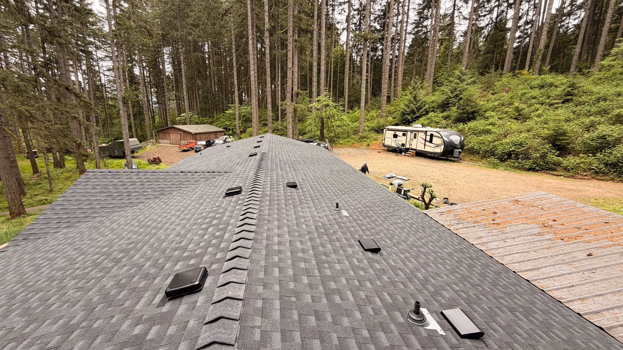 View along a new gray shingled roof with vents and surrounded by tall pine trees, an RV, vehicles, and a wooden shed nearby.