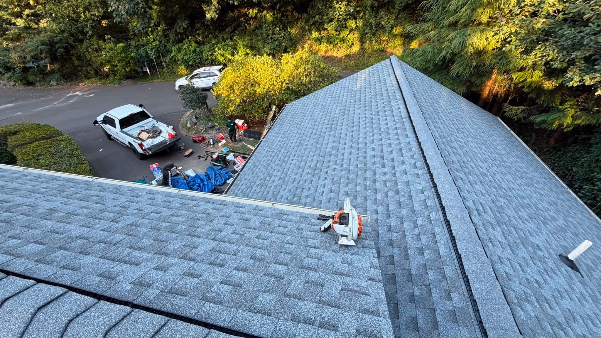 View from rooftop of gray shingles with tools, trucks, and workers below on driveway surrounded by trees.