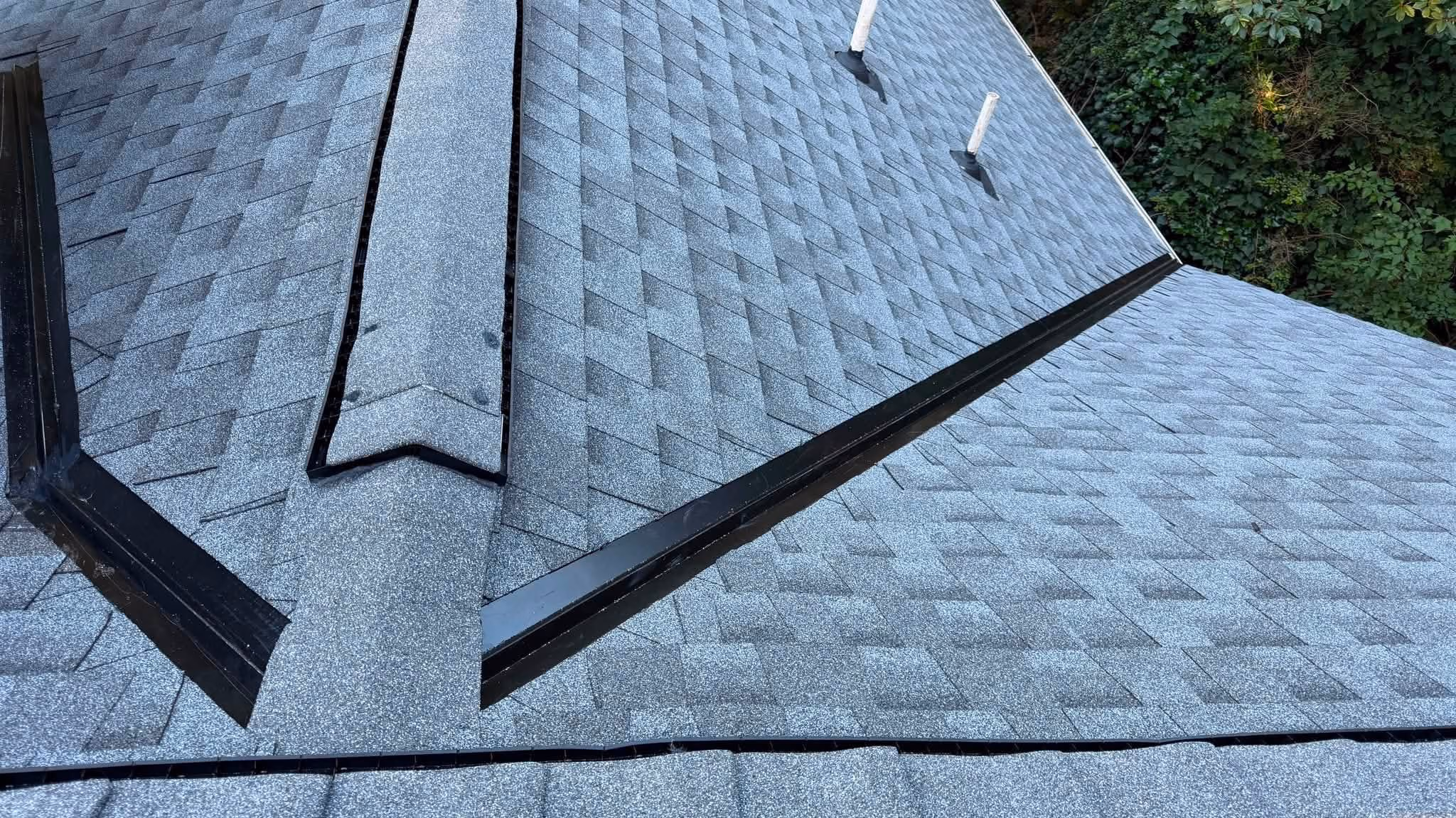 Gray shingled roof with ridge vents, flashing, and two white pipe vents surrounded by trees.