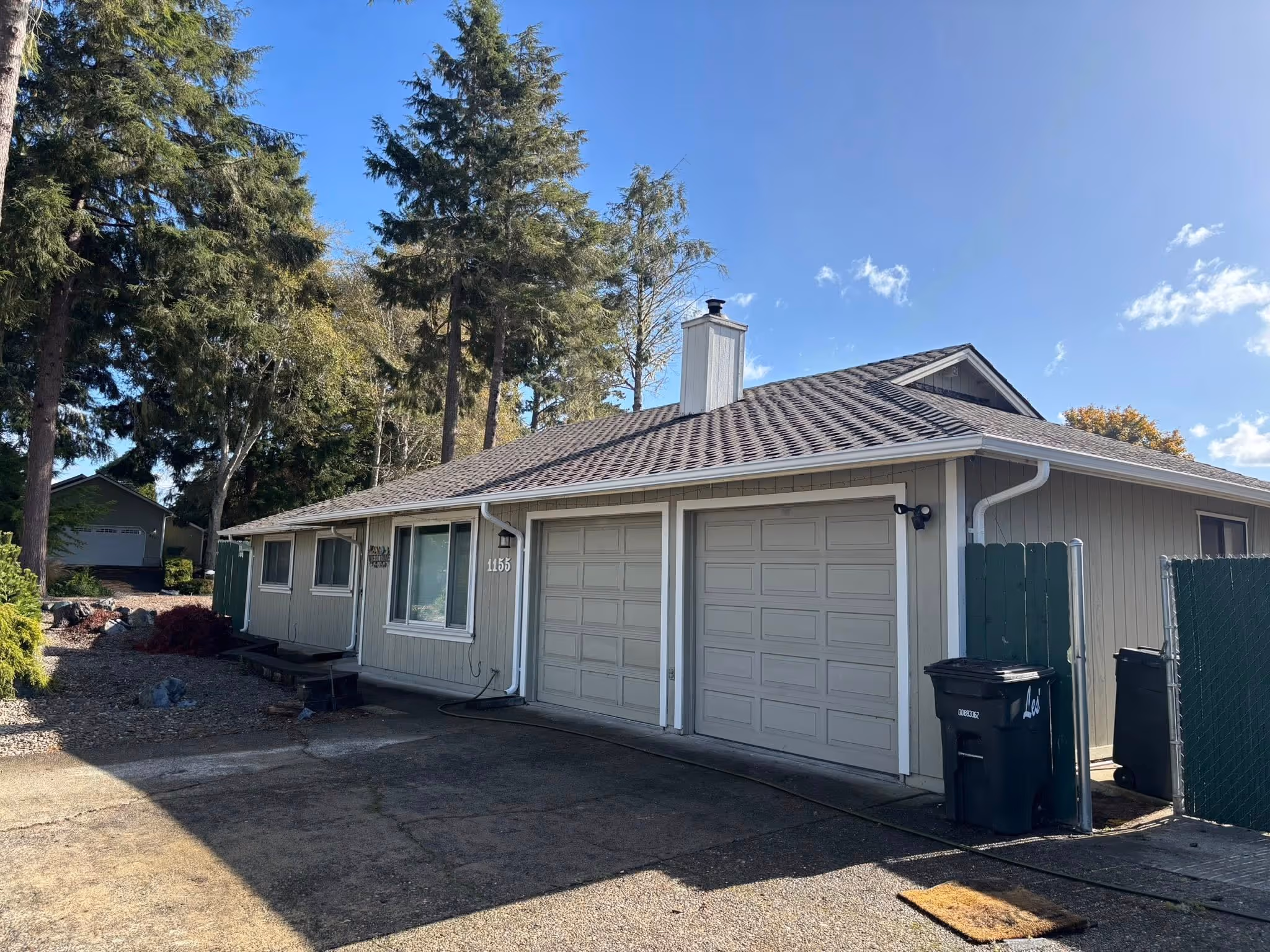 Single-story house with beige siding, two garage doors, surrounded by trees under a clear blue sky.
