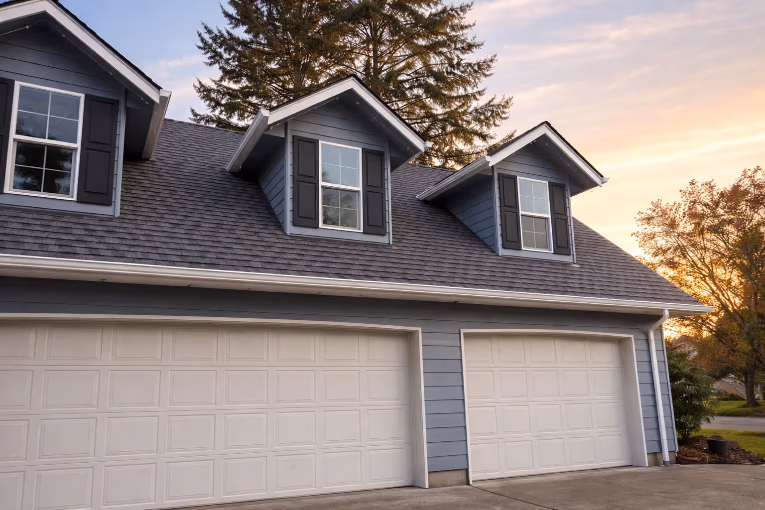 Blue house exterior with two white garage doors and three dormer windows under a dusky sky.