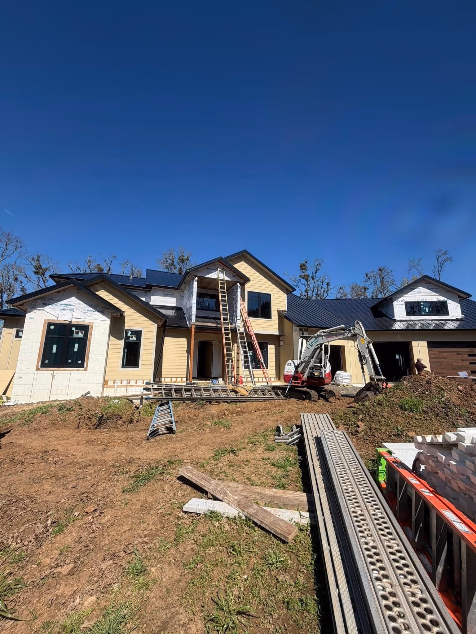 Partially constructed two-story house with yellow siding, black roof, ladders, construction equipment, and materials on a dirt yard under a clear blue sky.