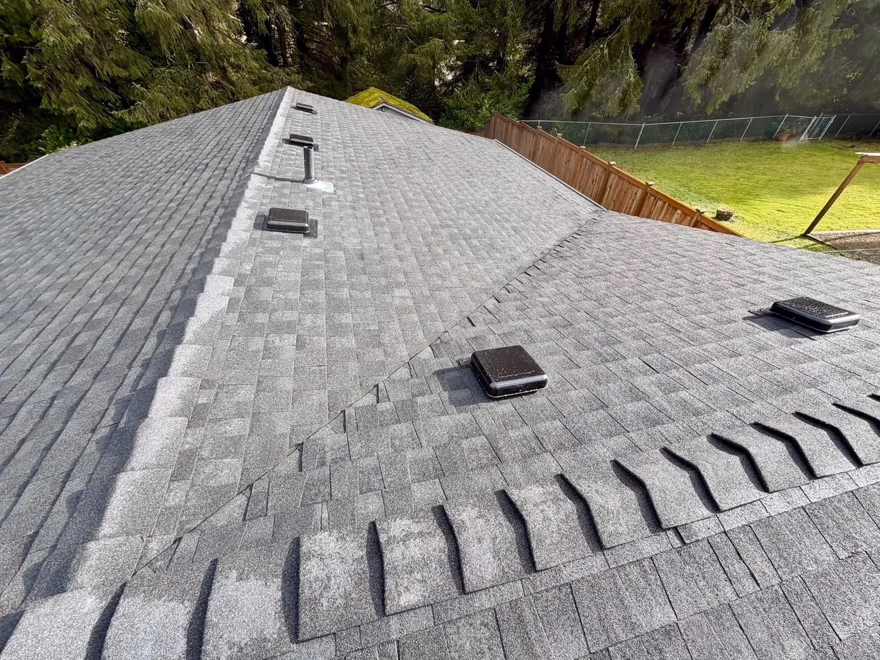 Gray asphalt shingle roof with multiple black vents and surrounded by trees and a wooden fence.