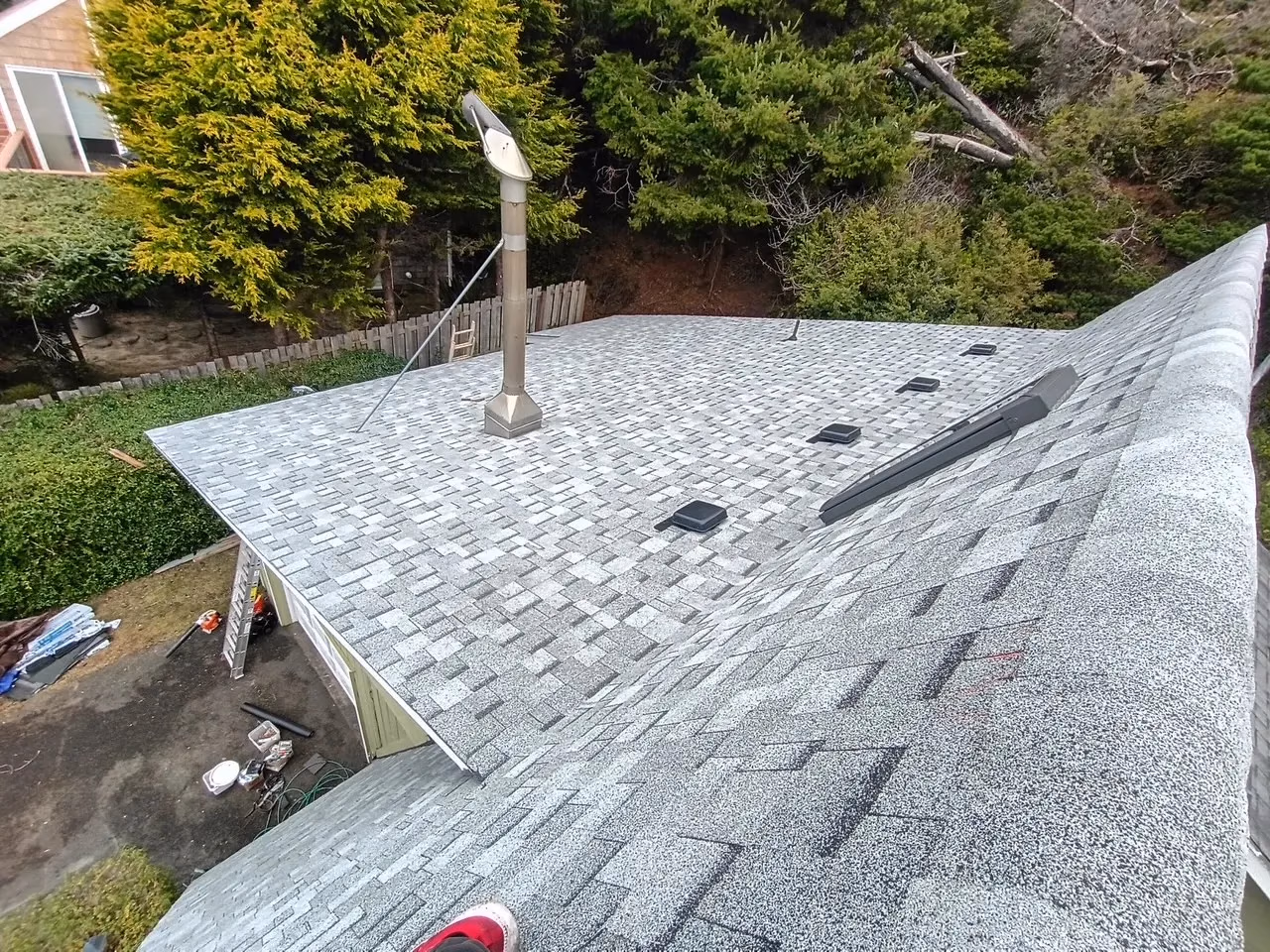 Gray asphalt shingle roof with multiple vents and a metal chimney pipe, surrounded by green trees.