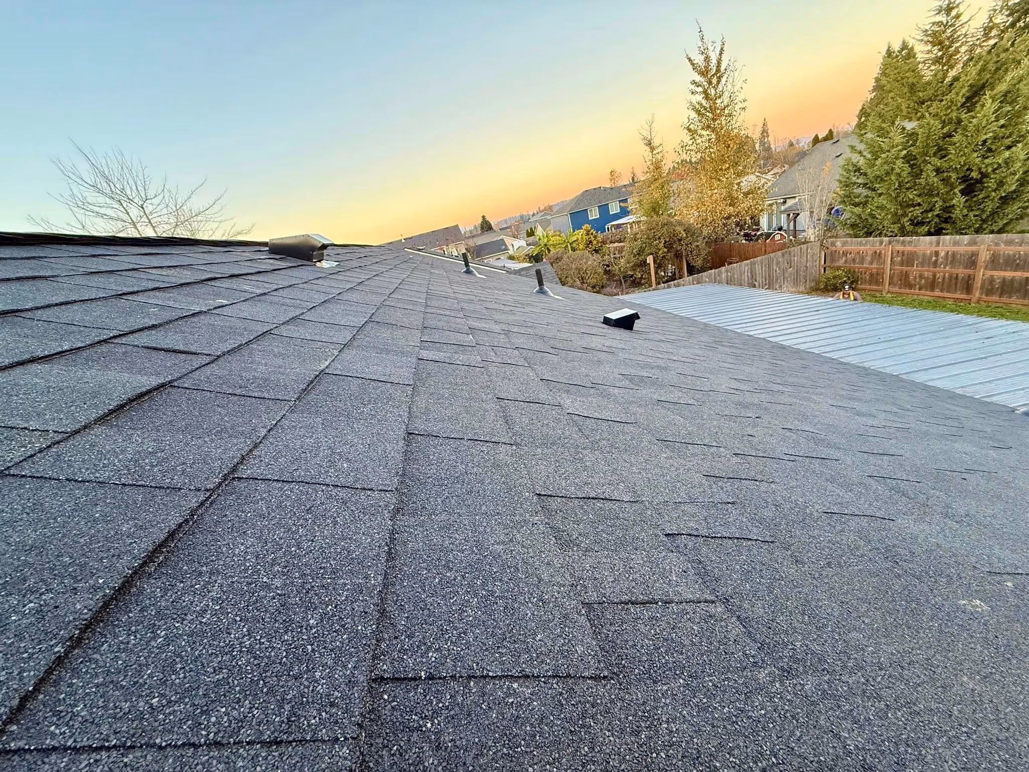 Close-up view of a gray asphalt shingle roof on a residential house at sunset with nearby trees and neighboring houses.