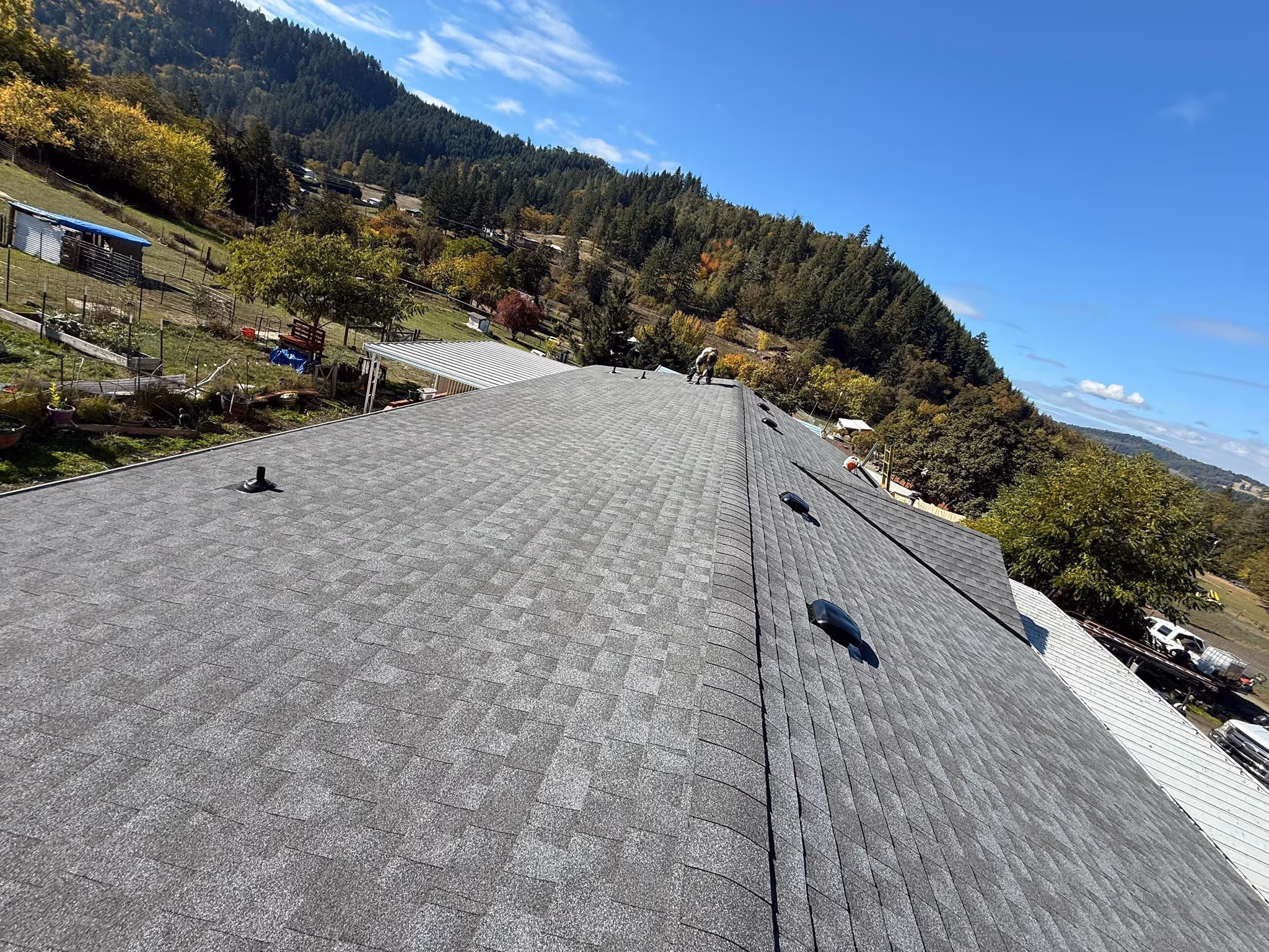 Gray shingled roof with vents and worker, surrounded by trees and hills under a clear blue sky.