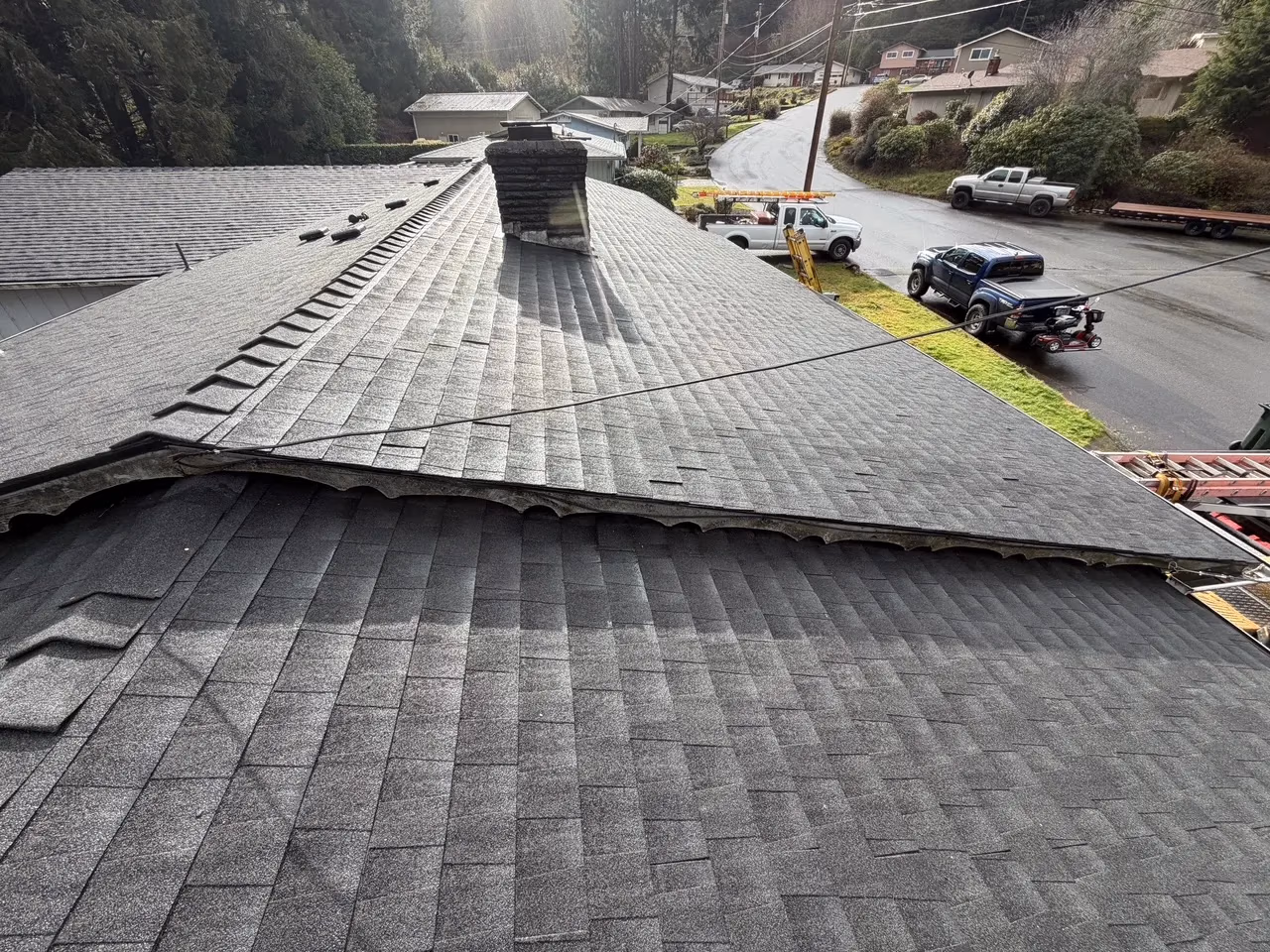 New grey asphalt shingle roof on a house with a chimney, surrounded by trees and several parked vehicles on the street.