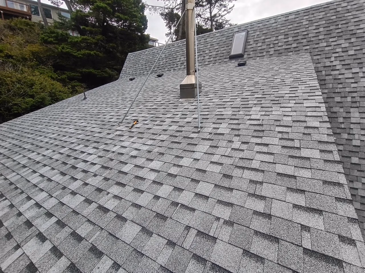 Grey asphalt shingle roof with a metal chimney and a small skylight, surrounded by trees.