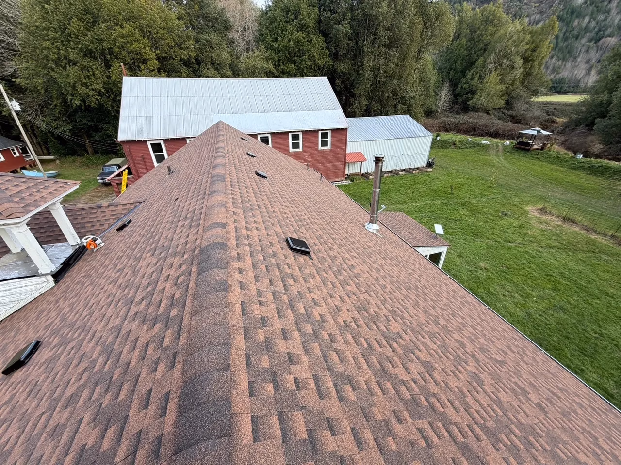 Bird's-eye view of a brown shingle roof with vents and a chimney, overlooking green lawn and trees with a red barn and a metal shed in the background.
