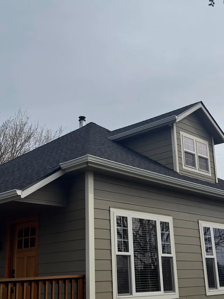 Side view of a two-story house with gray horizontal siding, white-framed windows, a black shingled roof, and a wooden front door partially visible under a covered porch.